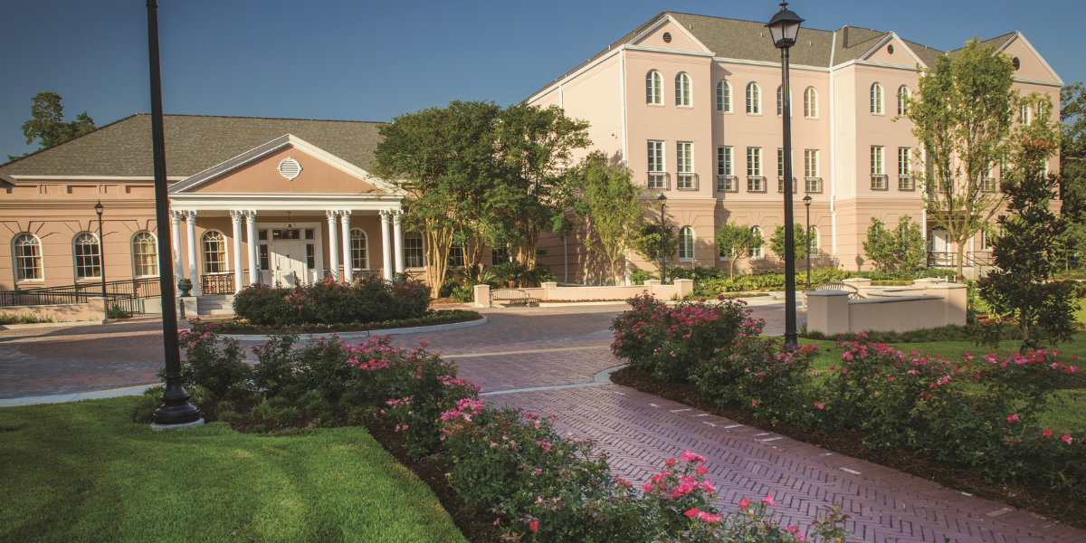 The beautifully landscaped entrance of Poydras Home in New Orleans, featuring pink rose gardens and a welcoming brick walkway leading to the memory care community.