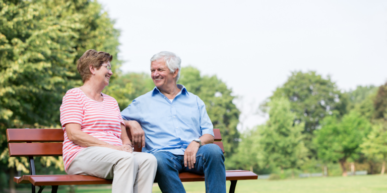 Senior couple smiling and talking together while sitting on a park bench outdoors.